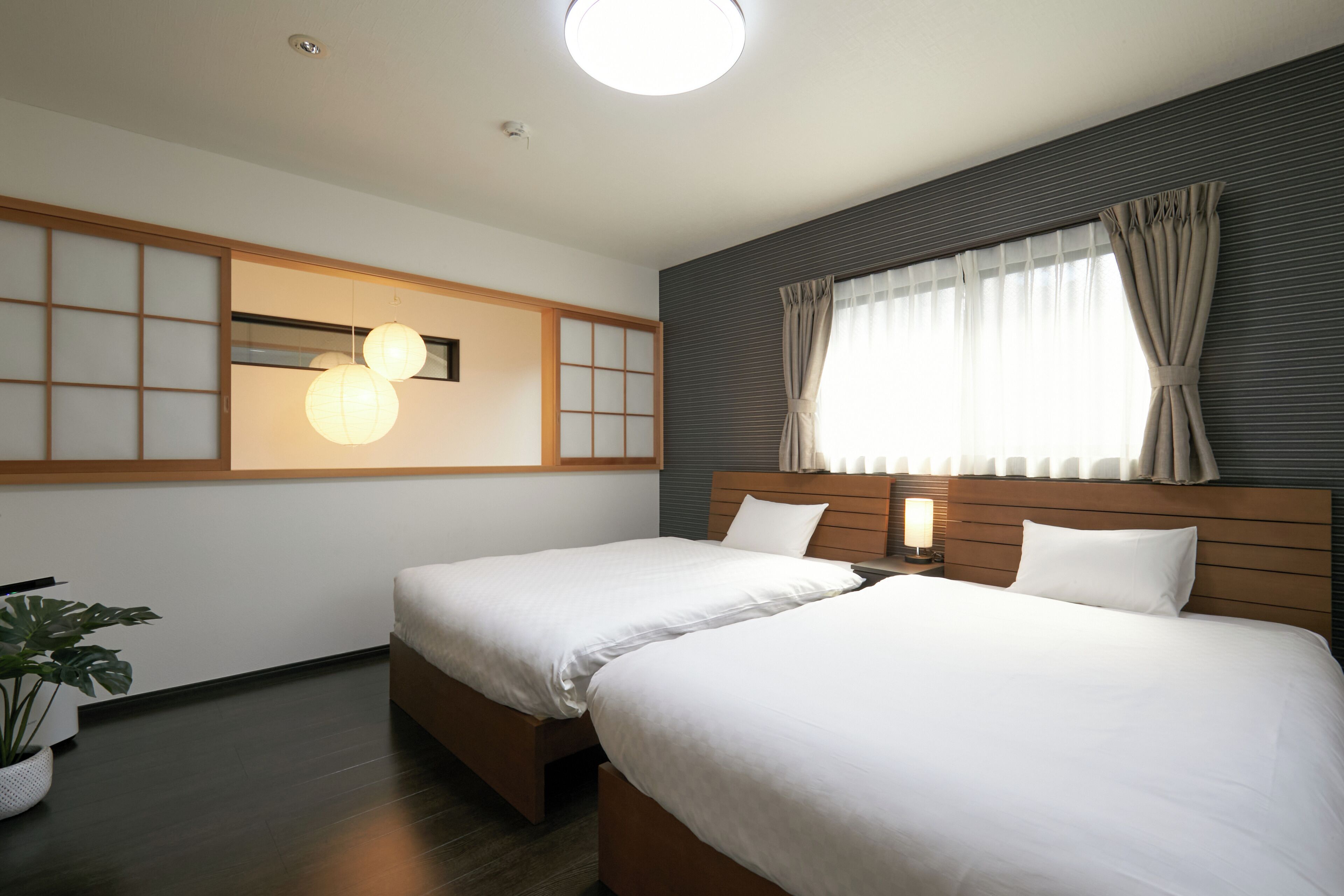 Une chambre d’hôtel avec deux lits, une grande fenêtre avec des rideaux, un luminaire fixé au mur et une plante en pot.