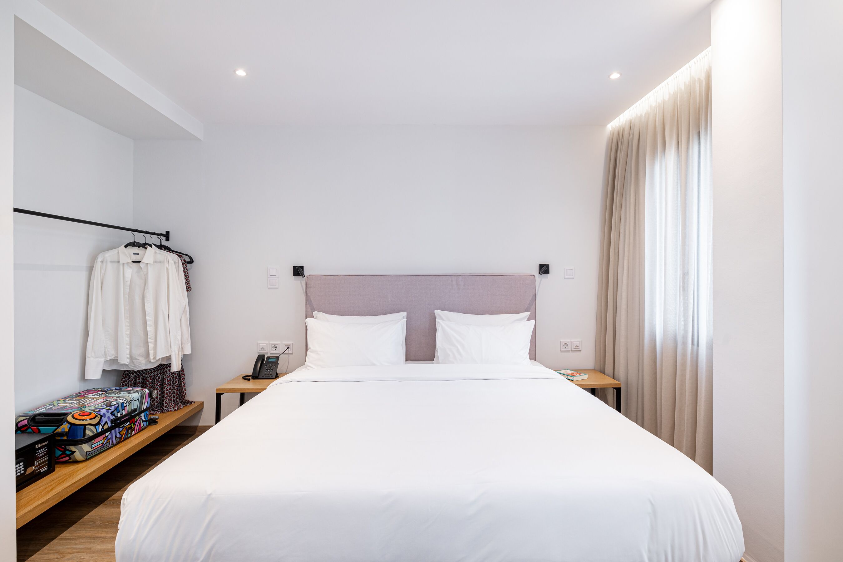 A neatly made bed with white linens, a wooden nightstand with a phone and a lamp, and a shelf with a suitcase and clothes.