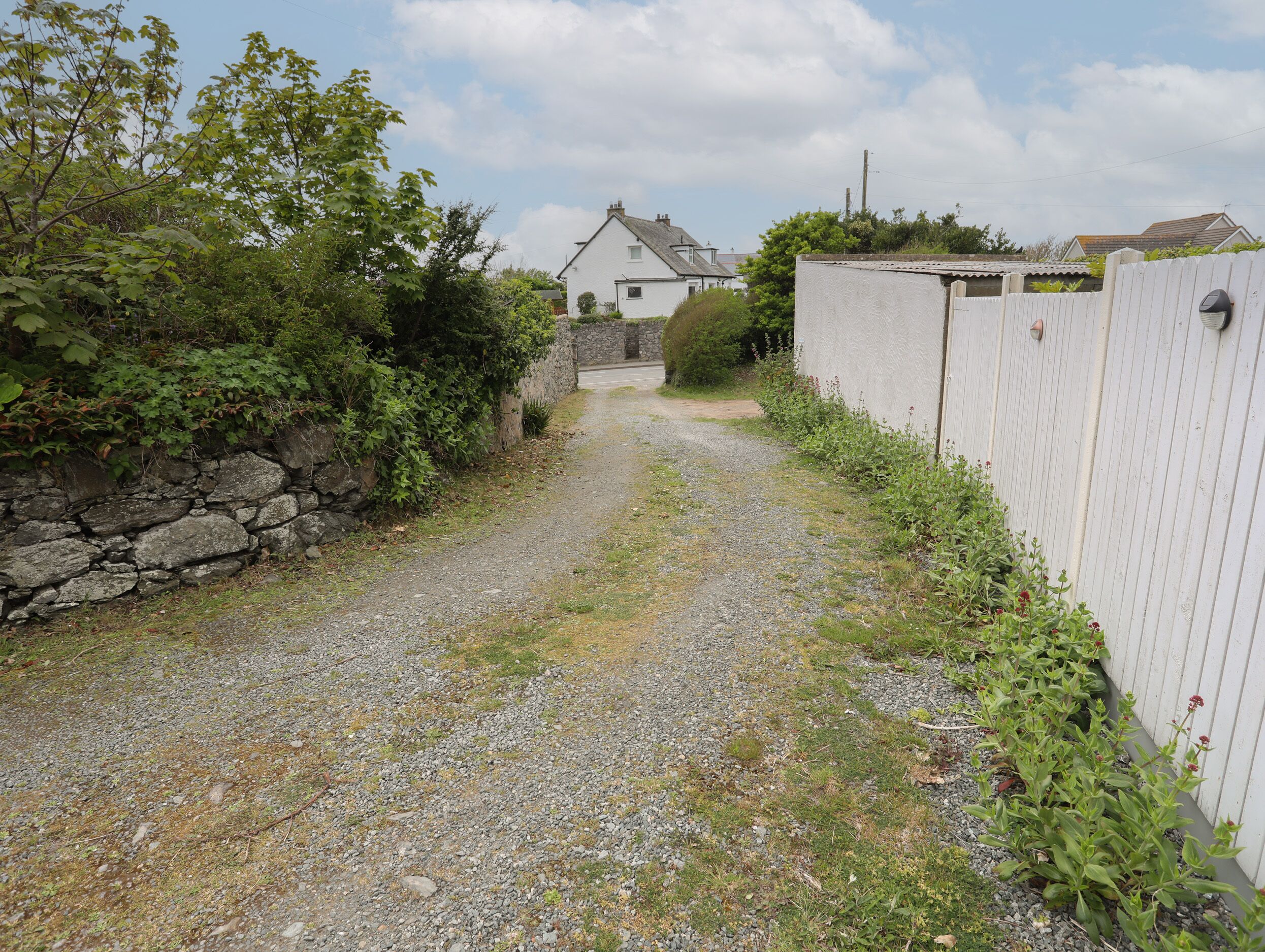Un chemin de gravier mène à une maison blanche, bordée d’un côté par un mur de pierres et de l’autre par une clôture blanche.