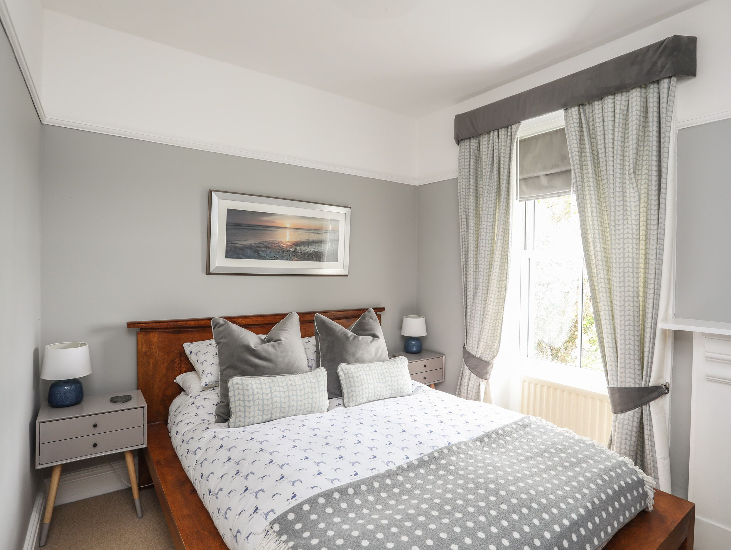 A bedroom with a bed, nightstands, a bedside table, a framed picture on the wall, and a window with curtains.