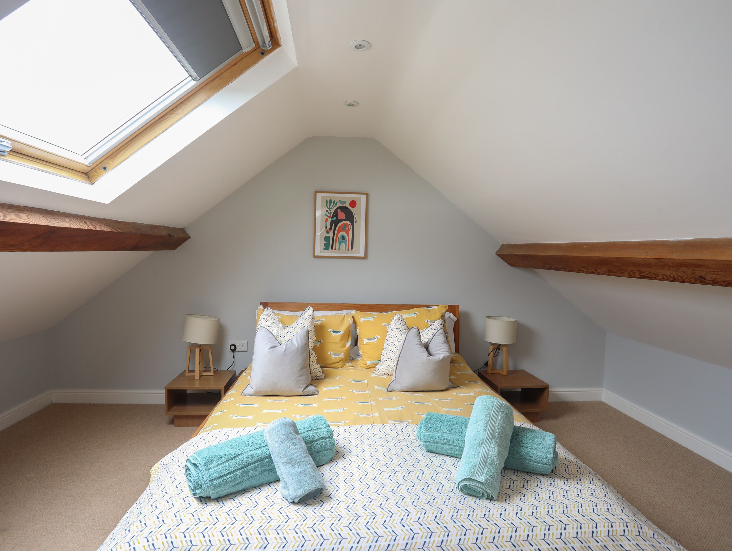A bedroom with a sloped ceiling, a skylight, a bed with patterned bedding, two bedside lamps, and a framed picture on the wall.
