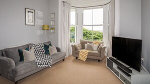 Un salon avec un canapé gris, un canapé beige, un téléviseur à écran plat et une grande fenêtre avec des rideaux.