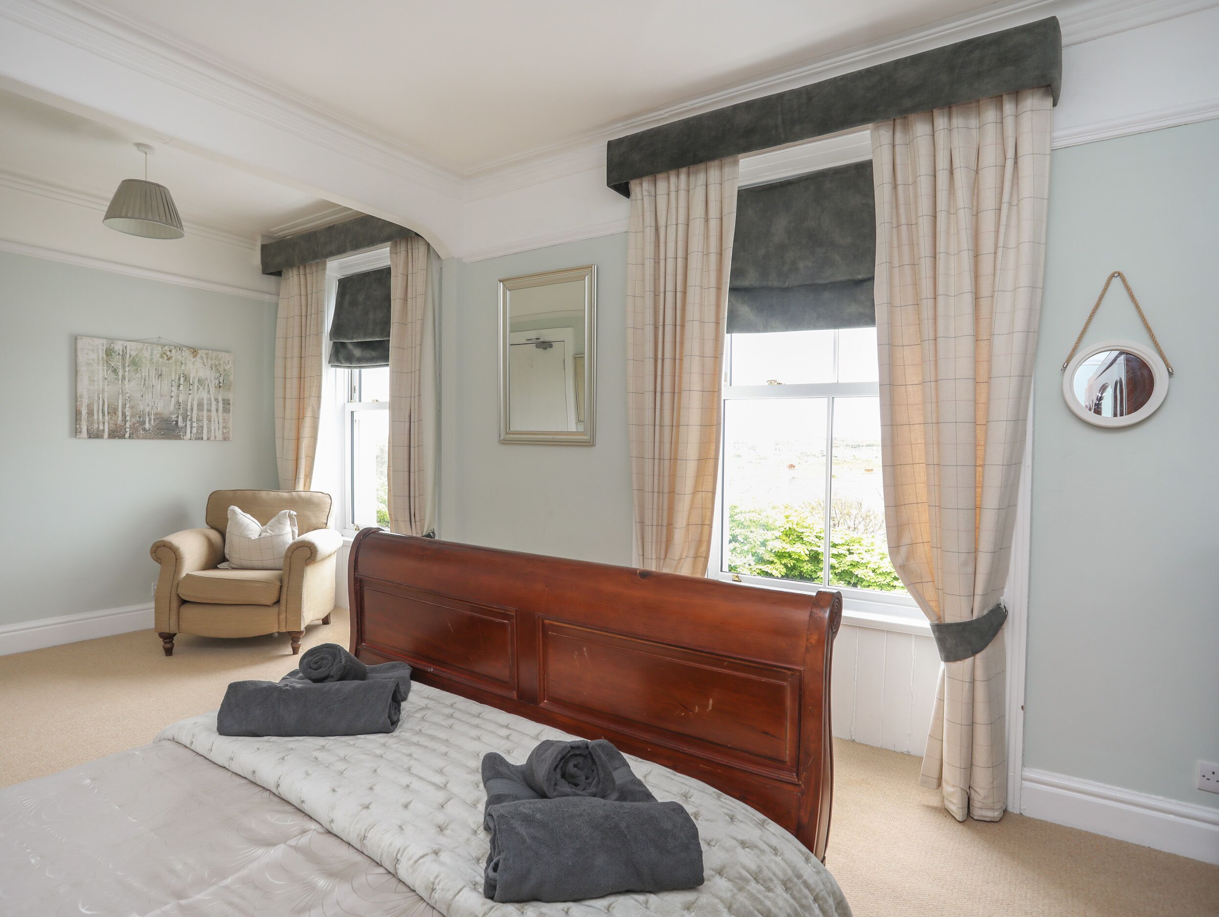A bedroom with a large bed, a chair, a window with curtains, a mirror, and a painting on the wall.