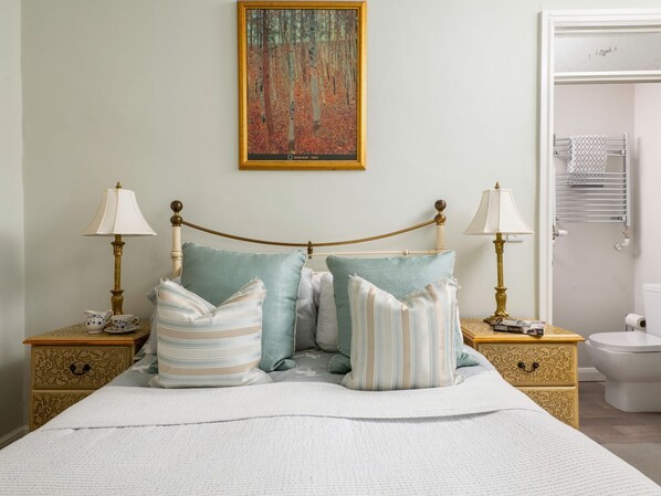Une chambre à coucher bien rangée, avec un lit, deux tables de chevet, un tableau, et une salle de bain visible à travers une porte ouverte.