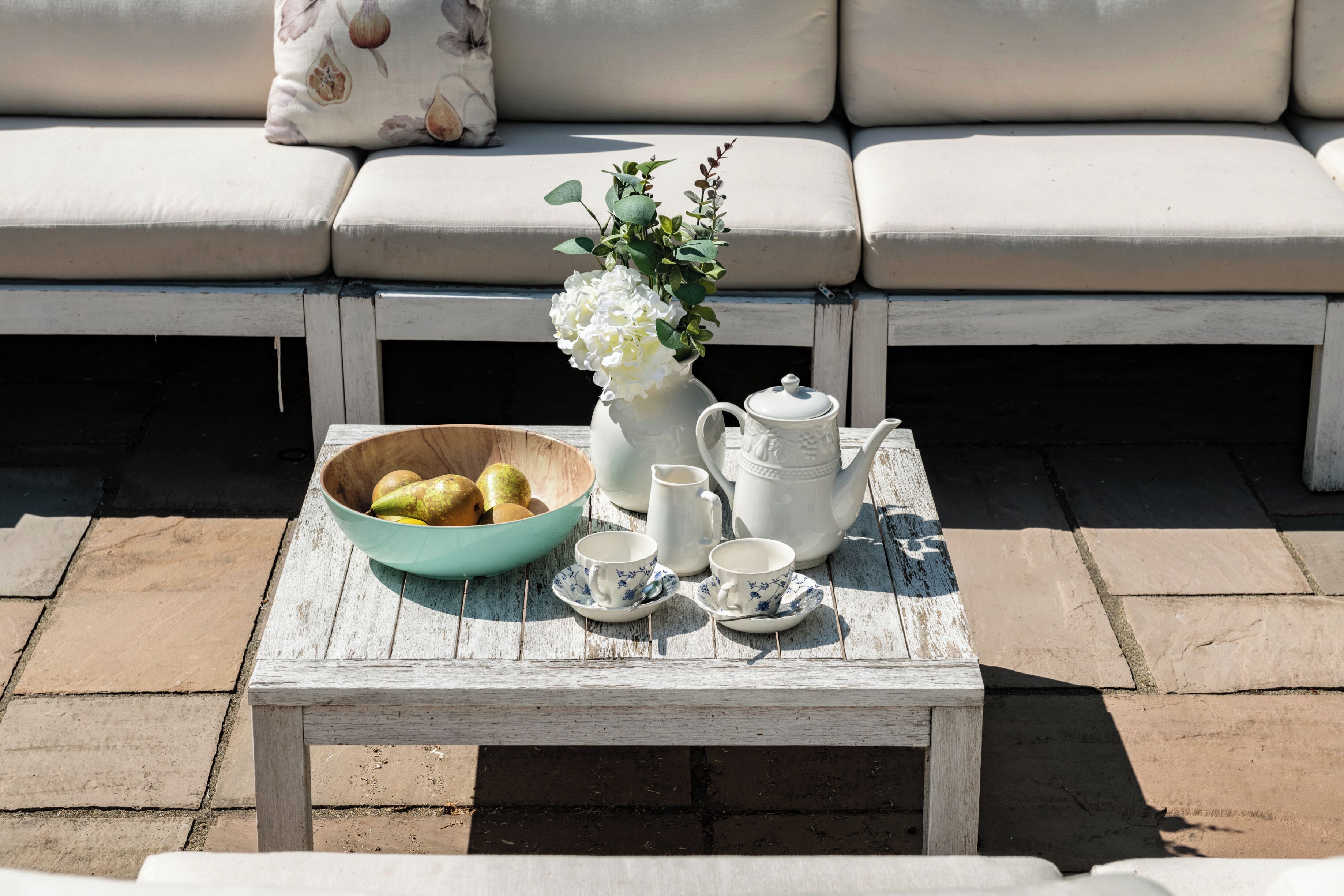Ein weißer Holztisch mit Obstschale, Blumenvasse, Teekanne und Tassen auf einer Terrasse, im Hintergrund ein weißes Sofa.