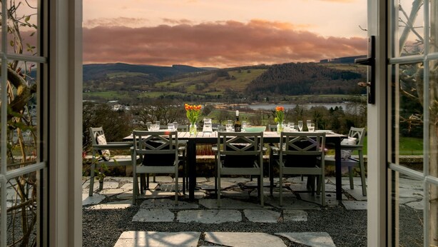 An open patio with a table set for two, overlooking a scenic landscape with hills and a body of water. - Fron Dderw (Bala)