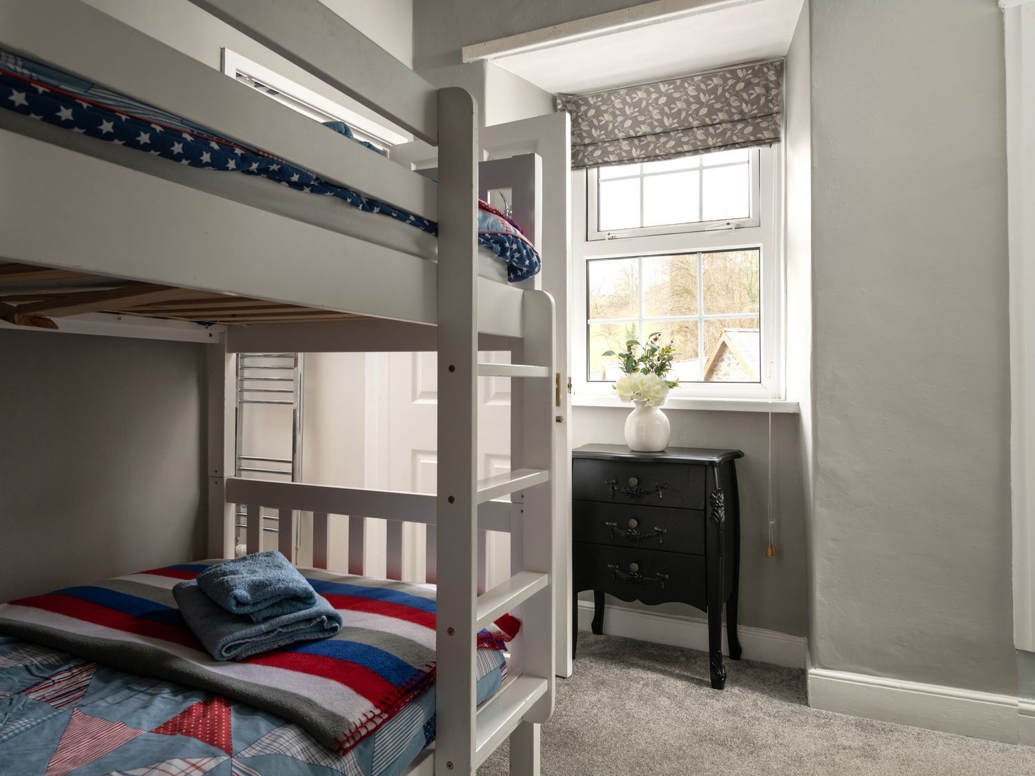 A bunk bed with a striped blanket, a small dresser with a vase of flowers, and a window with a view of trees.