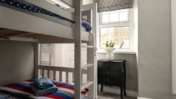 A bunk bed with a striped blanket, a small dresser with a vase of flowers, and a window with a view of trees.