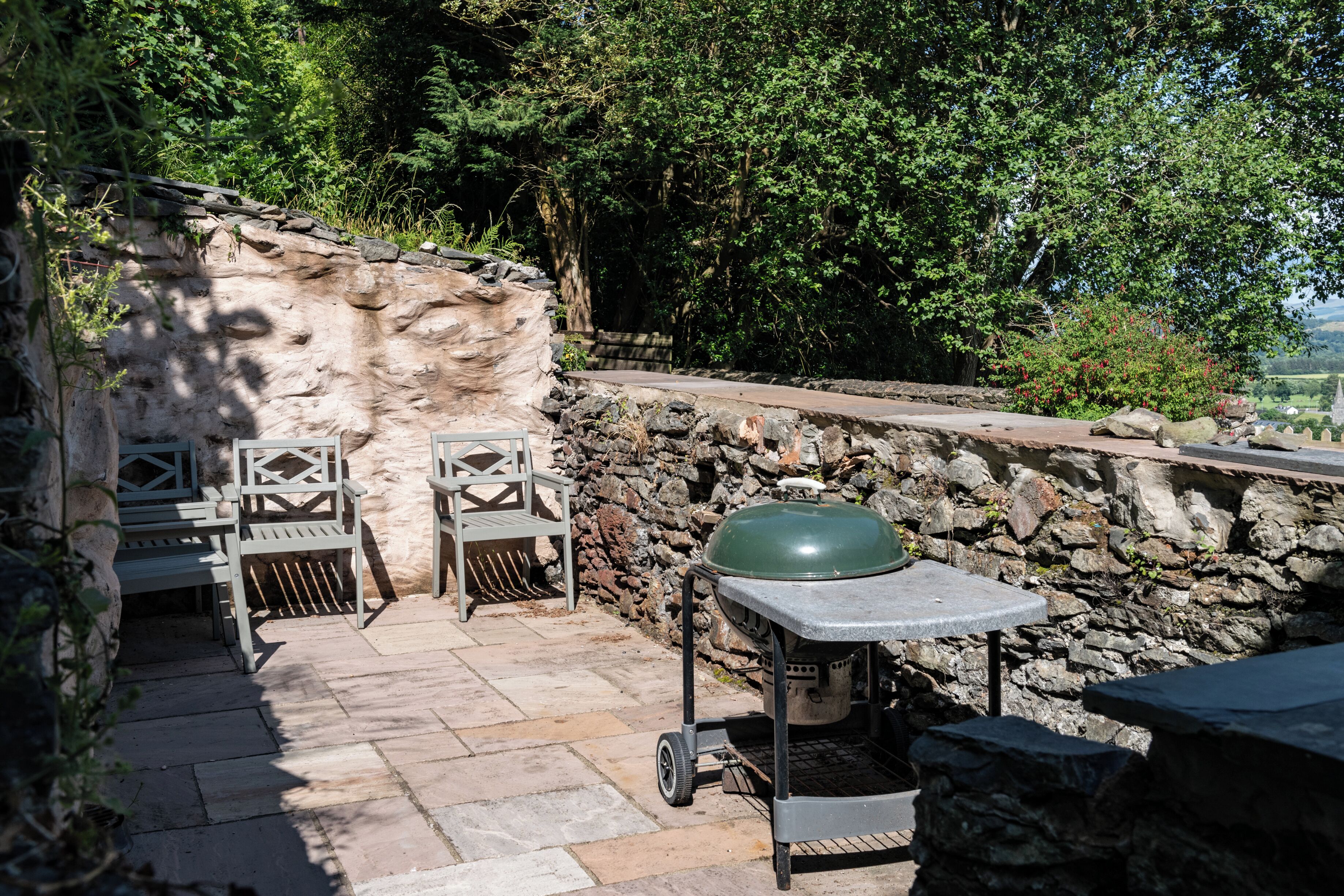 A stone wall, two white chairs, a green barbecue grill, and a stone table in a shaded outdoor area.