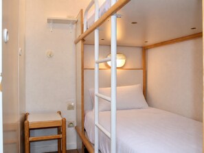 A bunk bed with a wooden frame and white bedding, a small wooden chair, and a wall-mounted shelf.
