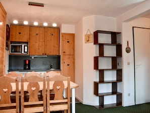 A kitchen with wooden cabinets, a microwave, and a coffee maker. There is a dining table with chairs and a wall-mounted shelf.