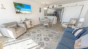 A living room with a sofa, a dining area with a table and chairs, a kitchen in the background, and a large TV on the wall. - Amazing Beachfront 1Bedroom at Bay Watch with Pool (North Myrtle Beach)