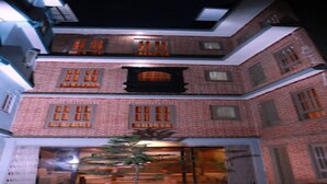 A multi-story building with a brick facade, large windows, and a visible entrance with a door and a small tree in front. - MeroStay 220 Hotel CBR (Bhaktapur)