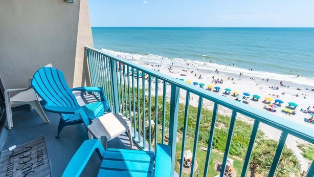 A balcony with blue chairs and a view of a beach with umbrellas.