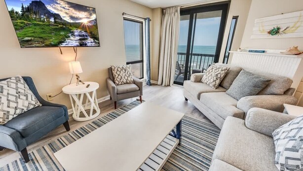 Ein modernes Wohnzimmer mit einer Couch, Sesseln, einem Couchtisch und einem an der Wand montierten Fernseher. Ein großes Fenster mit Vorhängen, ein Balkon mit Meerblick und ein Gemälde einer Berglandschaft.