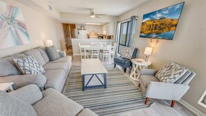 Ein modernes Wohnzimmer mit einem grauen Sofa, einem blauen Couchtisch und einem blauen Sessel. Im Hintergrund ist eine Essecke mit einem weißen Tisch und Stühlen zu sehen.
