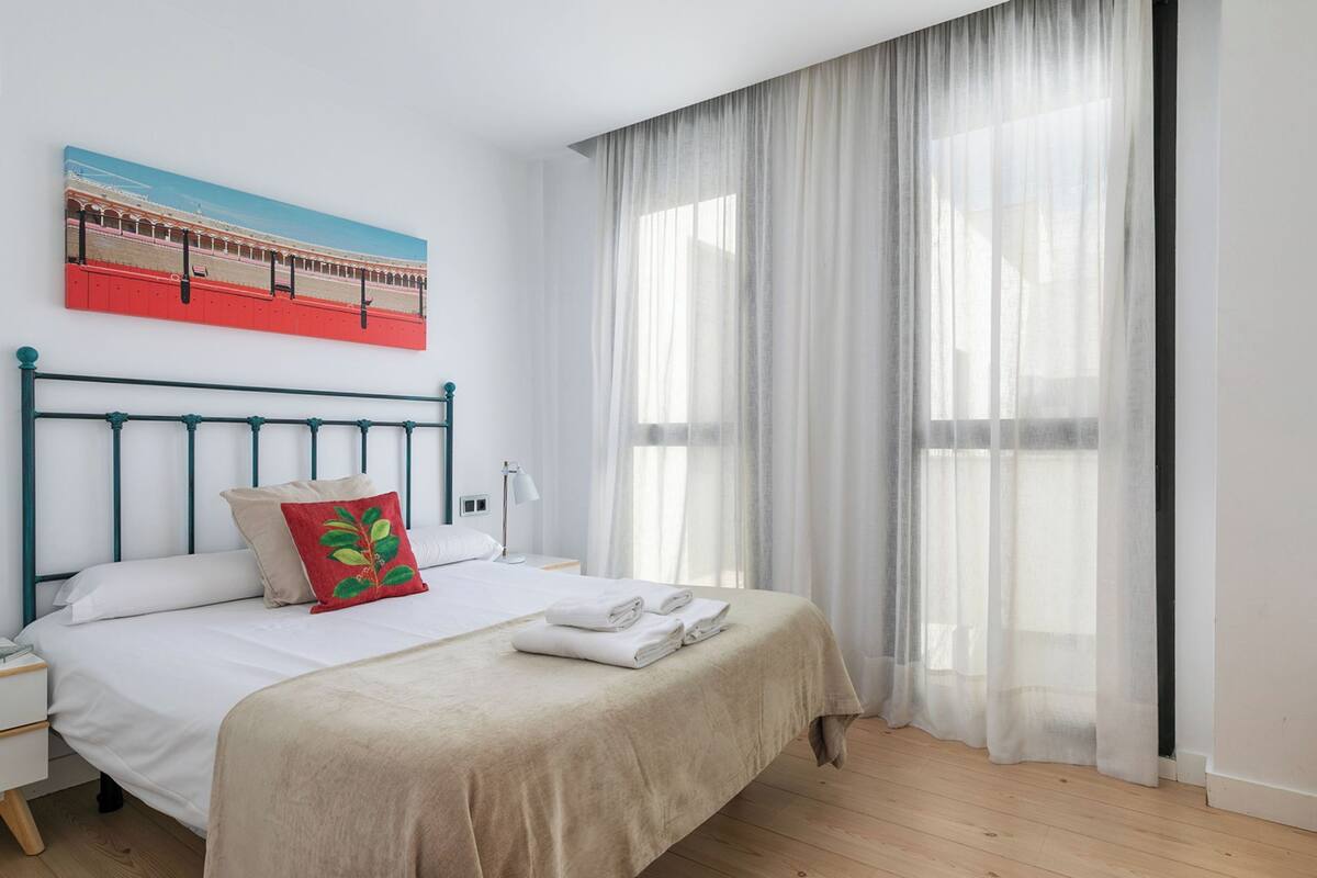 A neatly made bed with a beige bedspread and a red pillow, a white nightstand with a lamp, and a large window with sheer curtains.