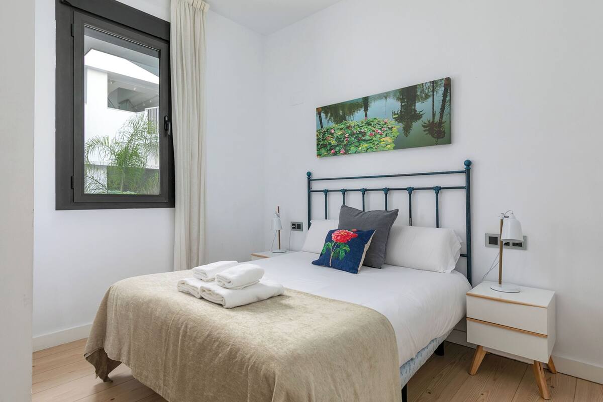 A neatly made bed with a beige bedspread and pillows, a bedside table with a lamp, and a window with curtains.