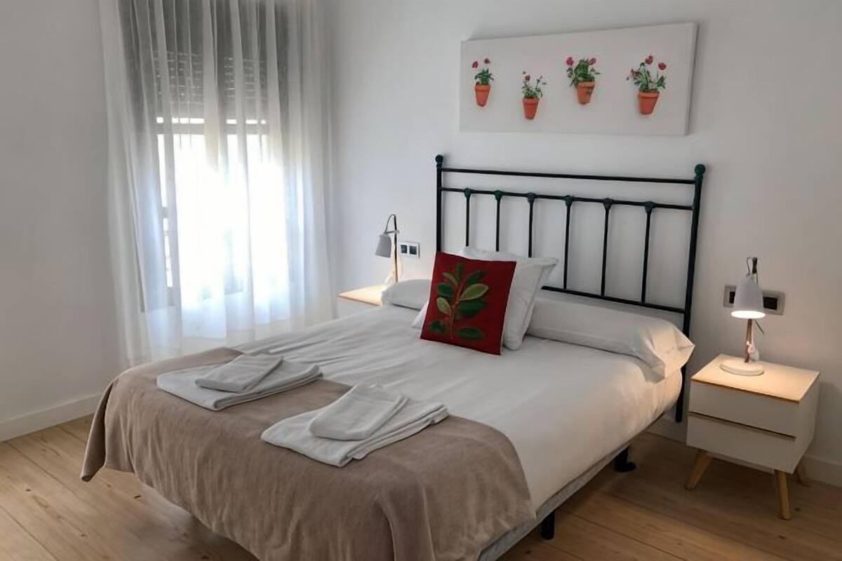 A neatly made bed with a headboard, two bedside tables with lamps, and a window with sheer curtains.