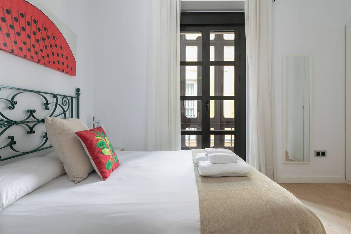 A bedroom with a bed, pillows, a red decorative pillow, a beige throw blanket, a mirror, and a door leading to an outdoor area.