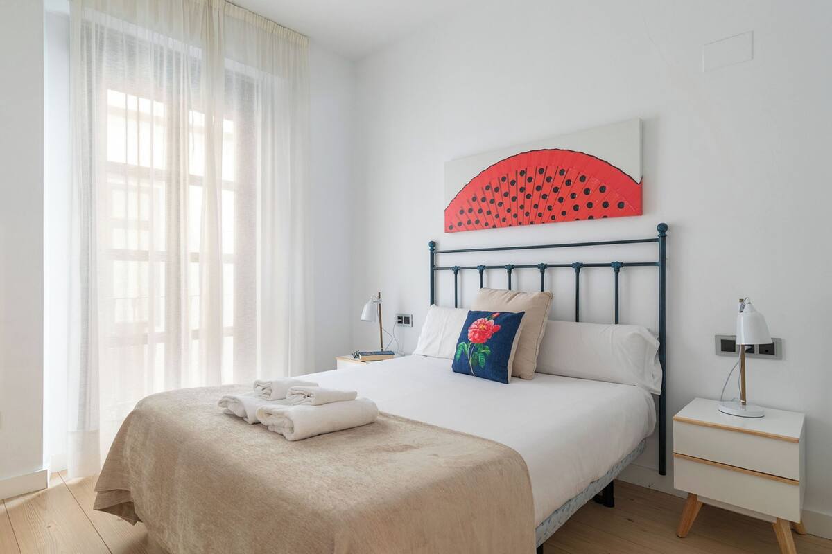 A neatly made bed with a beige bedspread, a red and white pillow, and a white pillow. There is a nightstand with a lamp and a small table beside the bed. A large window with sheer curtains allows natural light into the room.