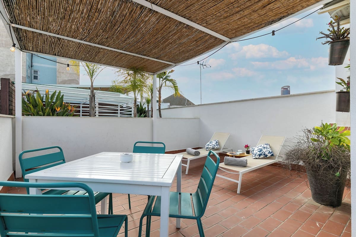 A rooftop terrace with a white table and chairs, a cushioned seating area, and potted plants.