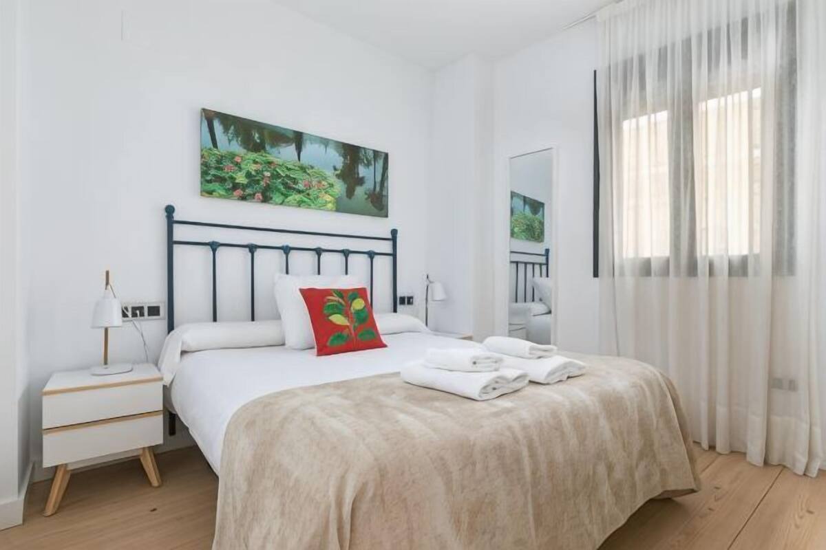 A bedroom with a bed, a nightstand, a bedside table, a window with curtains, and a painting on the wall.