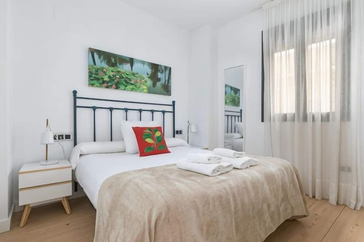 Um quarto com cama, mesa de cabeceira, cortinas na janela e um quadro na parede.