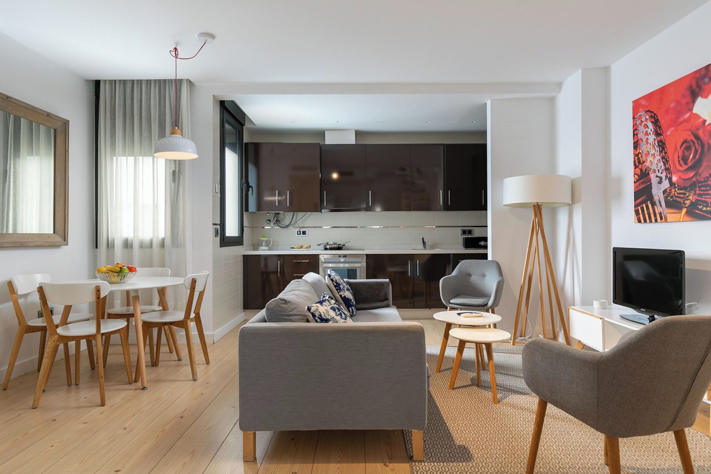 Een moderne woonkamer met een grijze bank, houten stoelen, een eettafel met fruit, een keuken op de achtergrond en een televisie op een dressoir.
