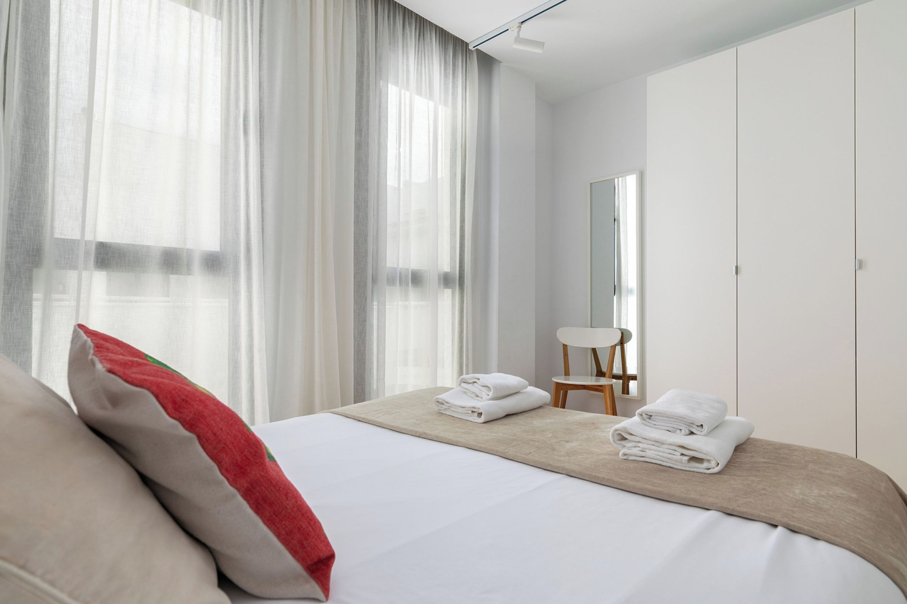 A modern hotel room with a neatly made bed, a small table with towels, a chair, and a large window with sheer curtains.