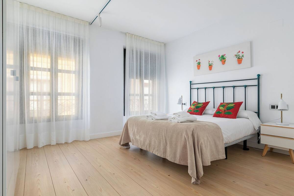 A bedroom with a bed, white curtains, a bedside table with a lamp, and a wall-mounted artwork of potted plants.