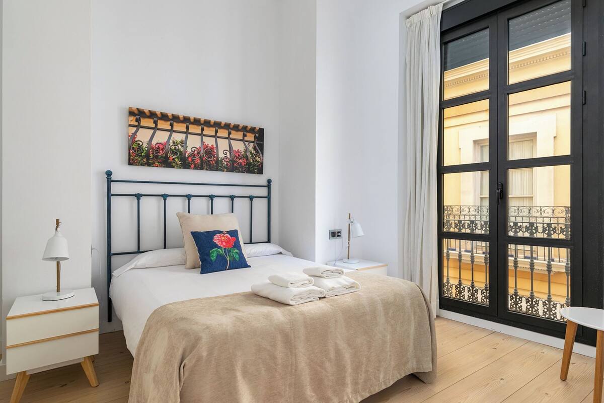 A bedroom with a bed, a nightstand, a lamp, a window with a view of a balcony, and a painting on the wall.