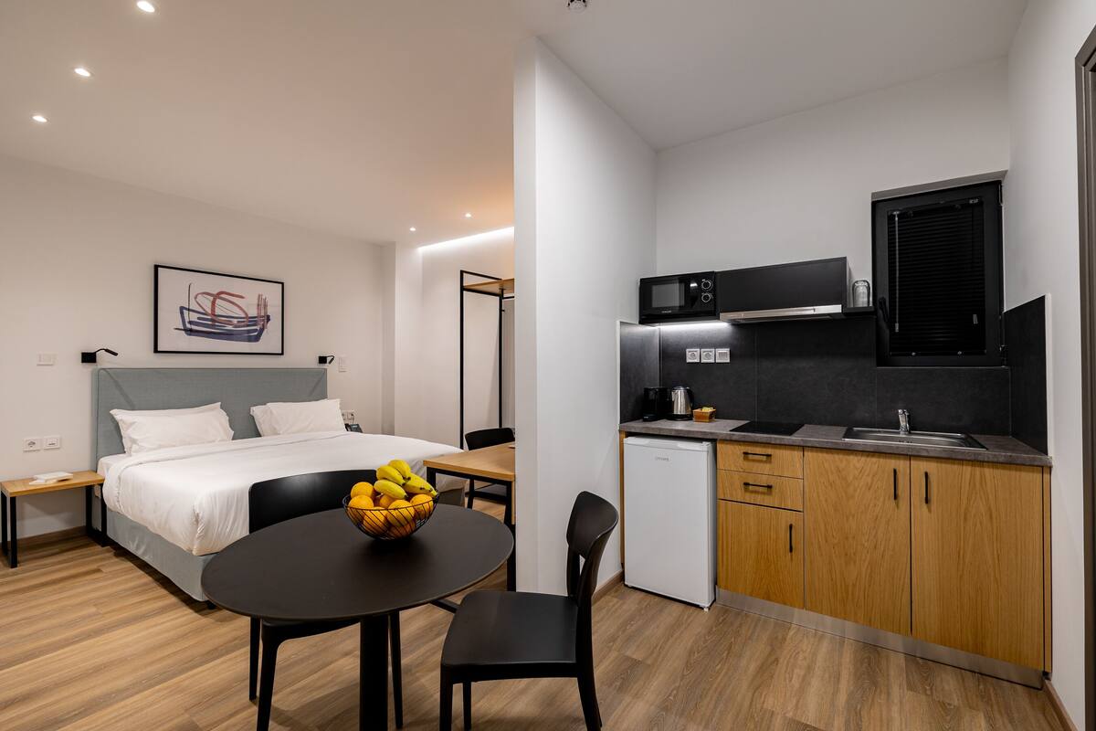 A compact hotel room with a bed, bedside table, a small dining table with chairs, a kitchen area with a microwave, sink, and cabinets, and a framed picture on the wall.