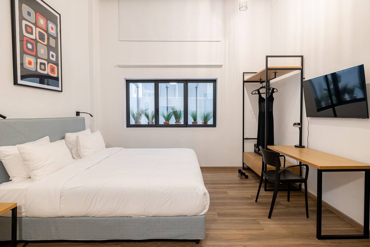 A modern bedroom with a bed, a desk, a chair, a TV, and a window with plants.