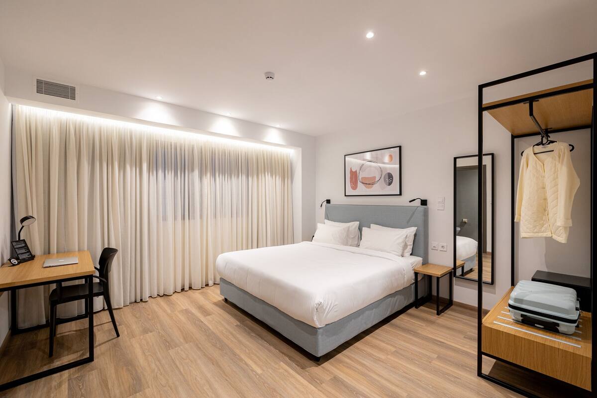A modern hotel room with a large bed, a desk, a chair, a wardrobe, and a mirror.