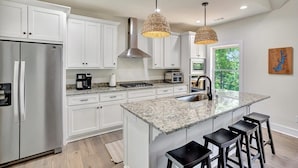 A modern kitchen with white cabinets, a stainless steel refrigerator, a granite countertop island with bar stools, and pendant lighting. - Peninsula Rd - Fun and Relaxation (West Union)