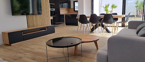 Una sala moderna con un televisor de pantalla plana, una mesa redonda y un sofá gris. Hay un comedor con una mesa y sillas de madera.