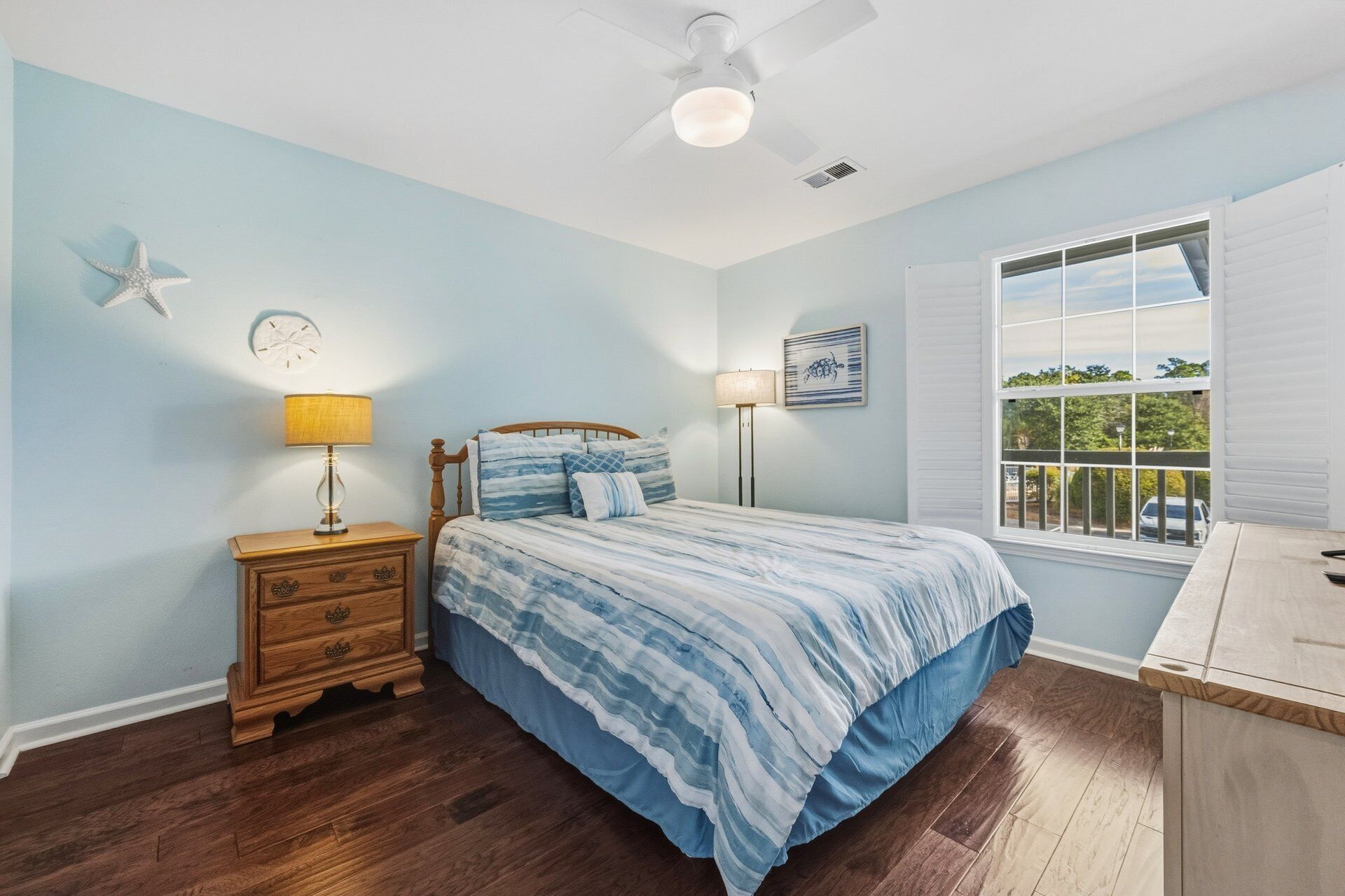 A bedroom with a bed, wooden nightstand, two wall-mounted lamps, a ceiling fan, a window with blinds, and a starfish decoration on the wall.