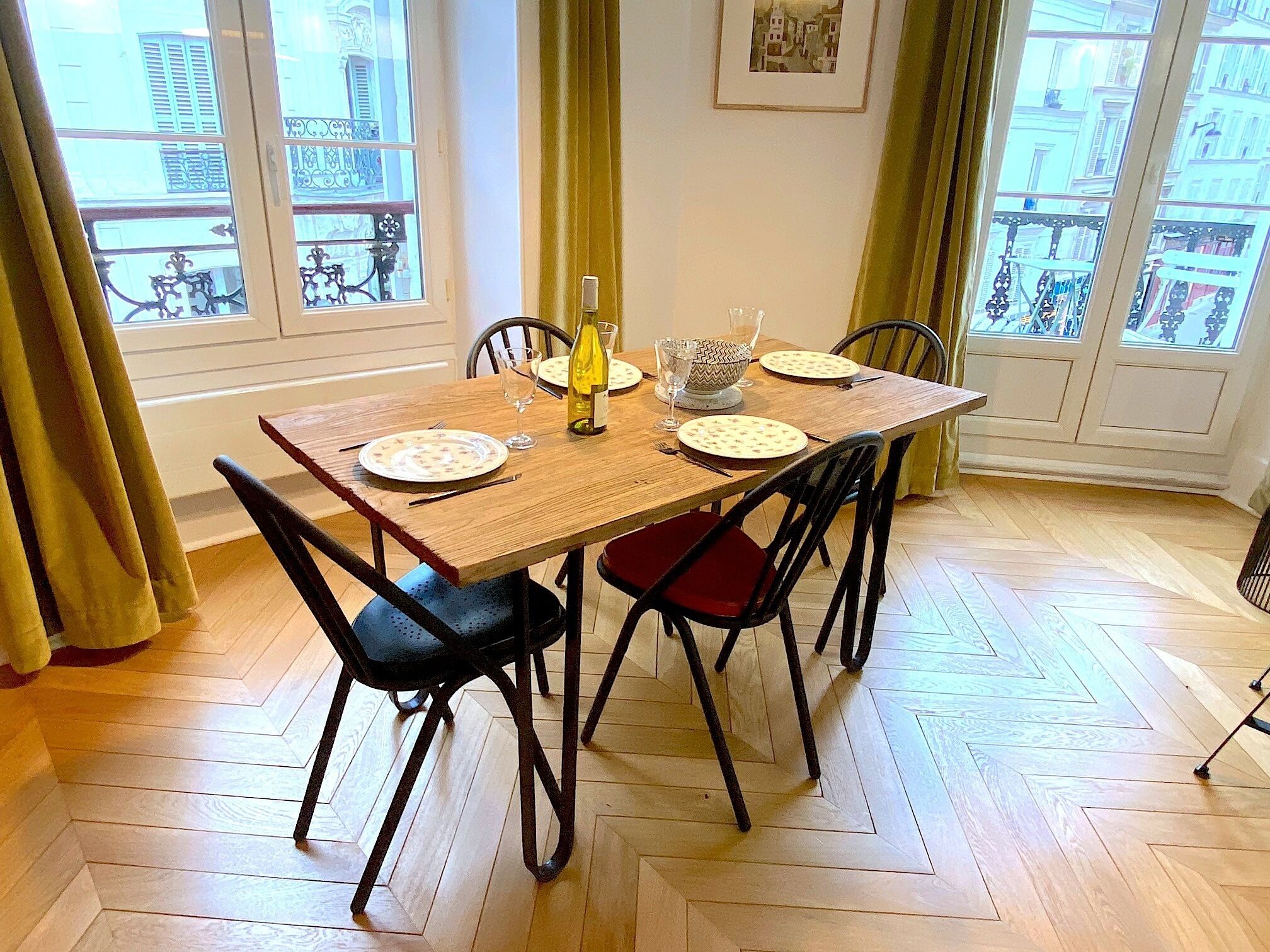 Una mesa de comedor con cuatro sillas, una botella de vino y un cuenco sobre un piso de madera con diseño de espiga.
