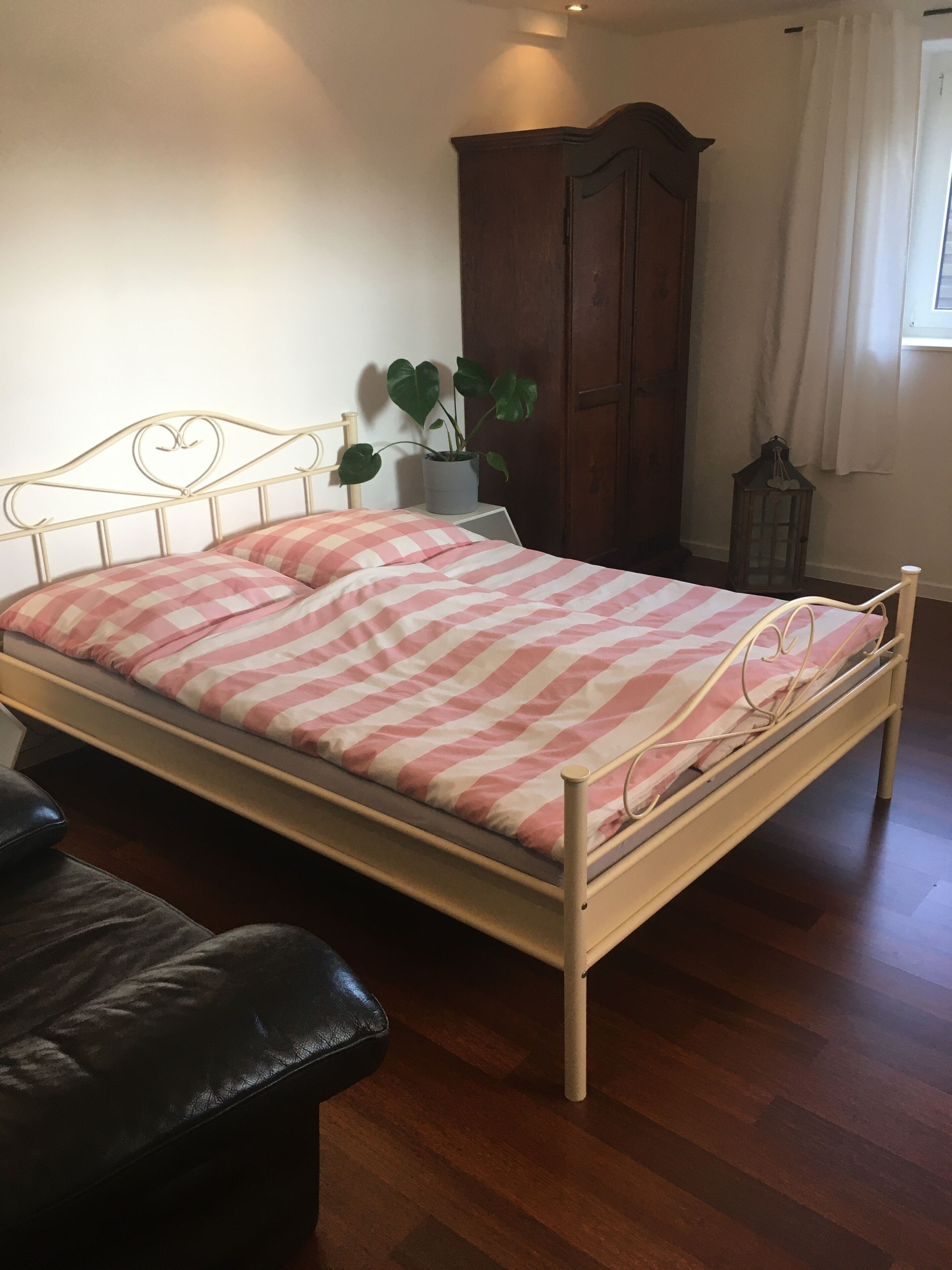 A bed with a striped bedspread, a wooden wardrobe, a potted plant, and a leather chair.