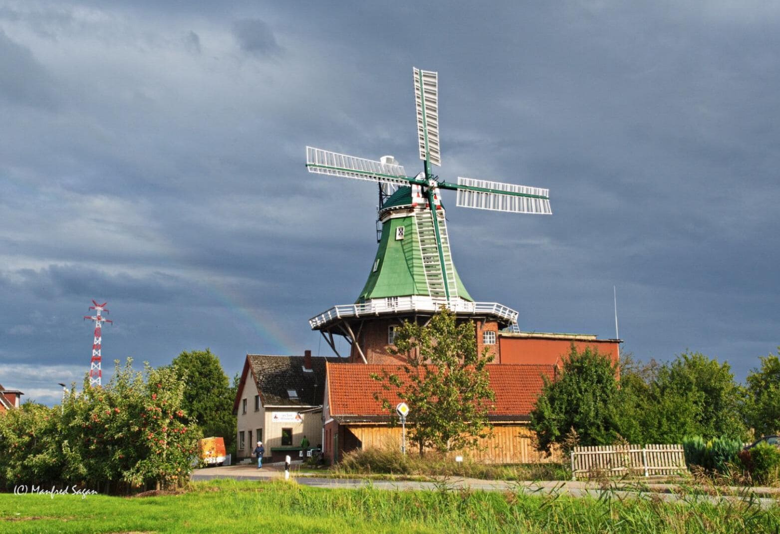 A traditional windmill with a green roof and white windmill blades, surrounded by greenery and a small building with a red roof.