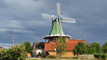 A traditional windmill with a green roof and white windmill blades, surrounded by greenery and a small building with a red roof.