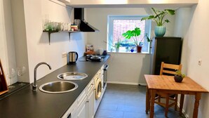Une cuisine compacte dotée d’un plan de travail noir, de deux éviers, d’une table à manger en bois et d’une fenêtre ornée de plantes.