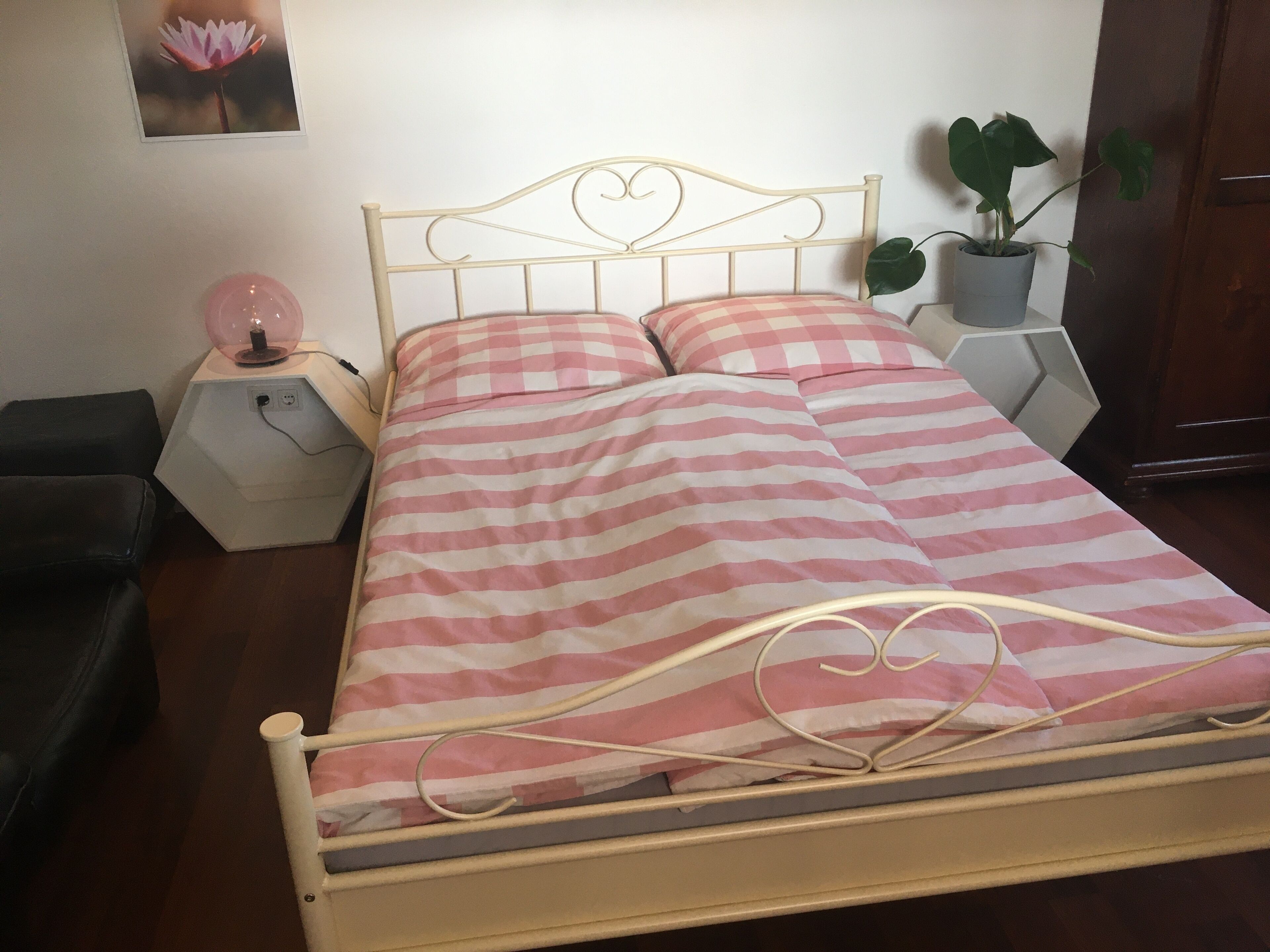 Una cama con estructura metálica y ropa de cama a rayas rosa y blanca, una lámpara de noche, una planta en maceta y un cuadro colgado en la pared.