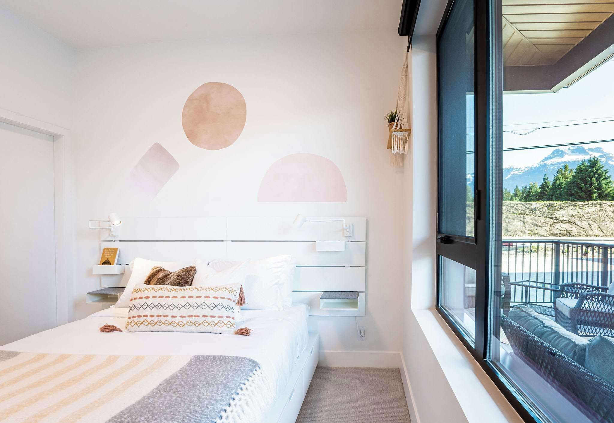 A bedroom with a bed, bedside tables, a window with a view of mountains, and a wall with geometric shapes.