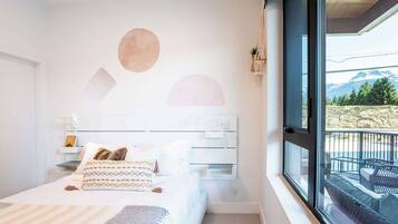 A bedroom with a bed, bedside tables, a window with a view of mountains, and a wall with geometric shapes.