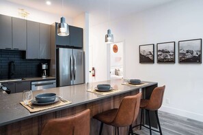 A modern kitchen with dark cabinets, a stainless steel refrigerator, and a central island with bar stools. - Summit Place - Condo With Mountain Views (Revelstoke)
