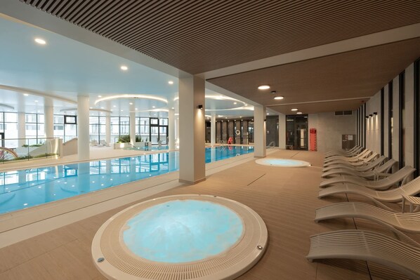 A modern indoor swimming pool with lounge chairs, a hot tub, and a well-lit ceiling.