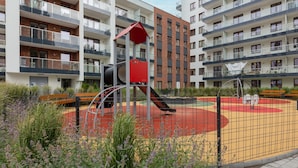 Un parque infantil rodeado por una valla, junto a un edificio residencial con balcones y una estructura roja para jugar.