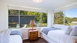 Une chambre avec deux lits, une table de chevet et une lampe. De grandes fenêtres offrent une vue sur une maison et sur la verdure environnante.